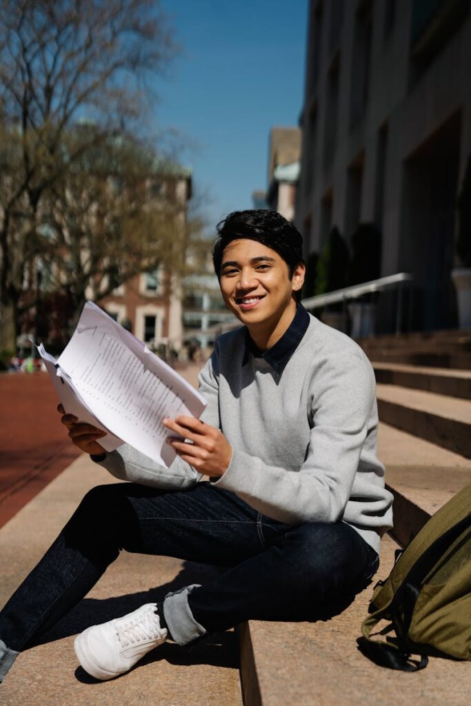 Young student smiling while holding papers on university campus steps.